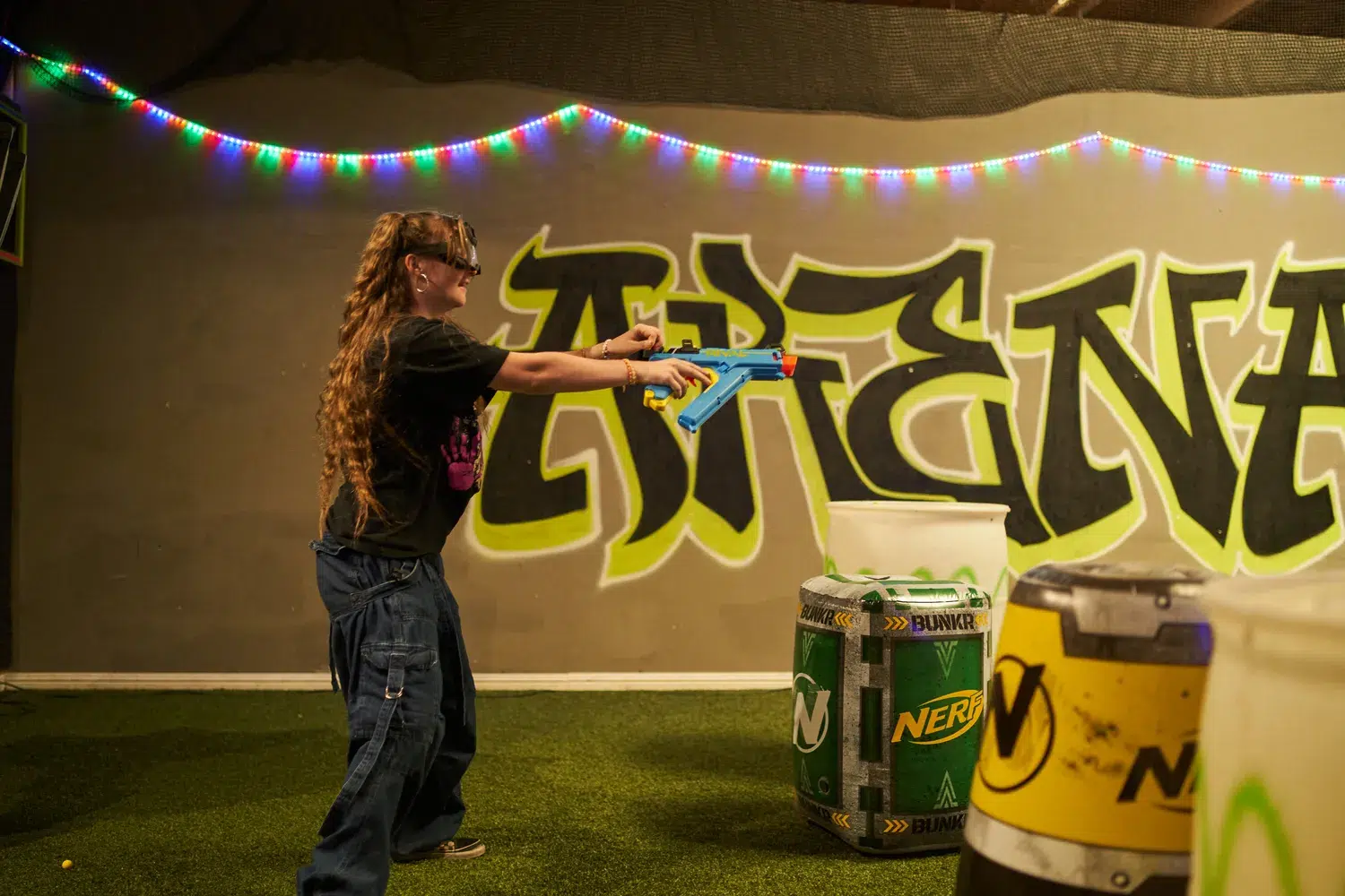 A girl safely engages in a Nerf battle at an indoor arena, equipped with protective eyewear. The vibrant setting, featuring graffiti and branded barriers, emphasizes fun and safety in action-packed play.