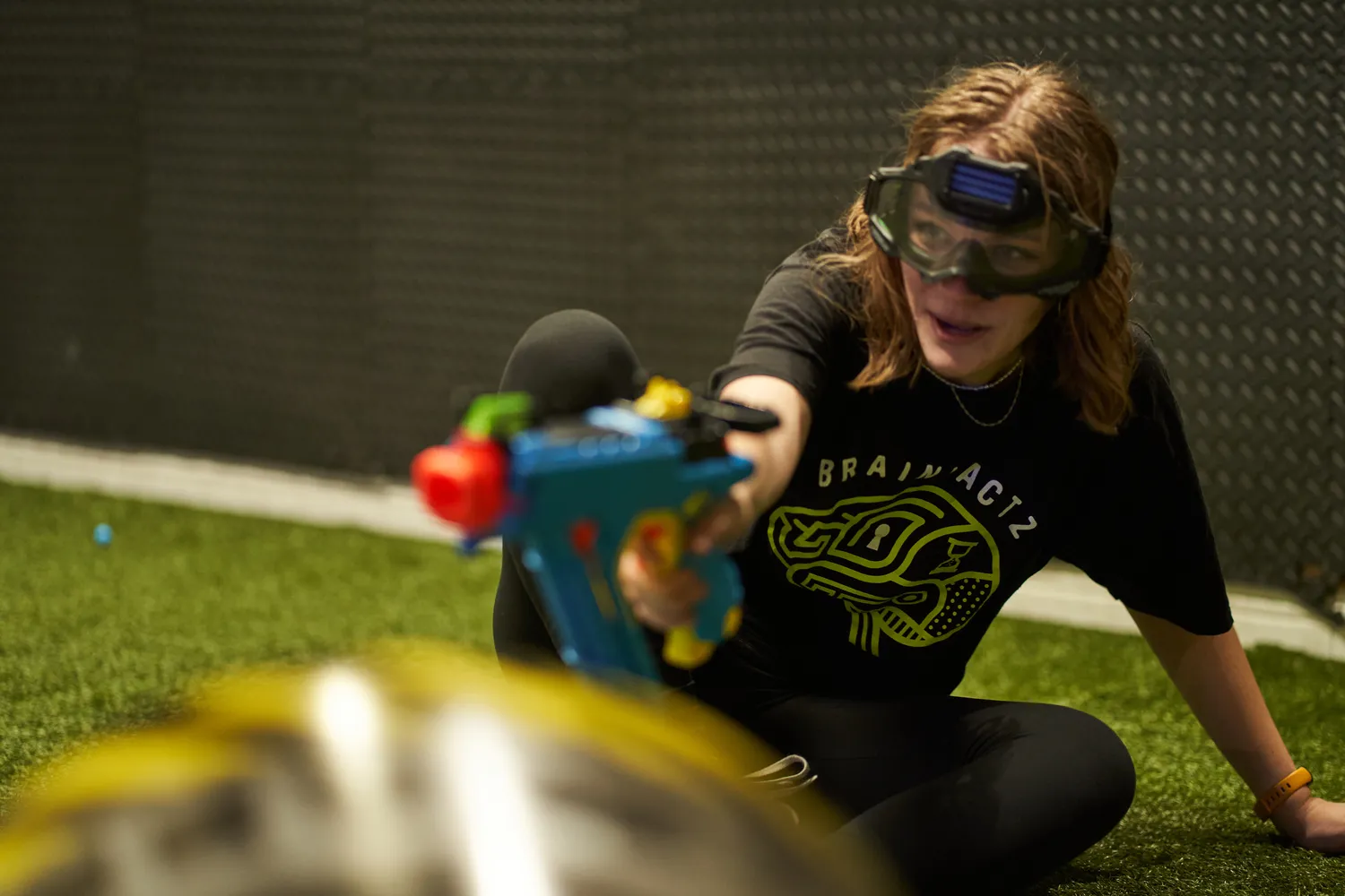  A person aiming a Nerf blaster while crouching, wearing protective eyewear, with foam darts scattered on the ground, demonstrating essential gear for Nerf competitions.