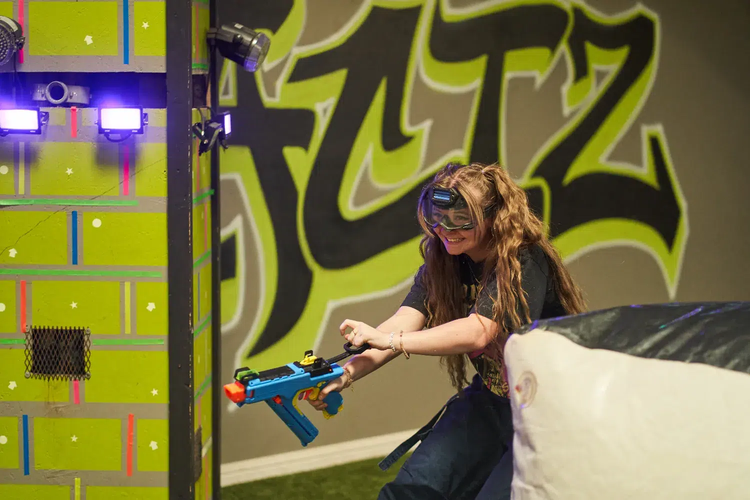 Smiling woman crouching and aiming a Nerf blaster in a colorful indoor play area.