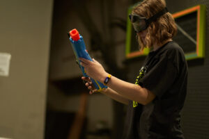 Copy of A7E01785 - Brainy Actz Escape Rooms Young player examining a blue Nerf blaster, focusing on its features to choose the best Nerf gun, while wearing safety goggles in an indoor gaming arena.