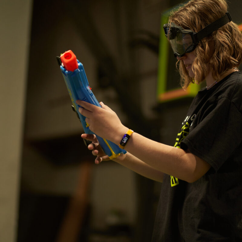 Young player examining a blue Nerf blaster, focusing on its features to choose the best Nerf gun, while wearing safety goggles in an indoor gaming arena.