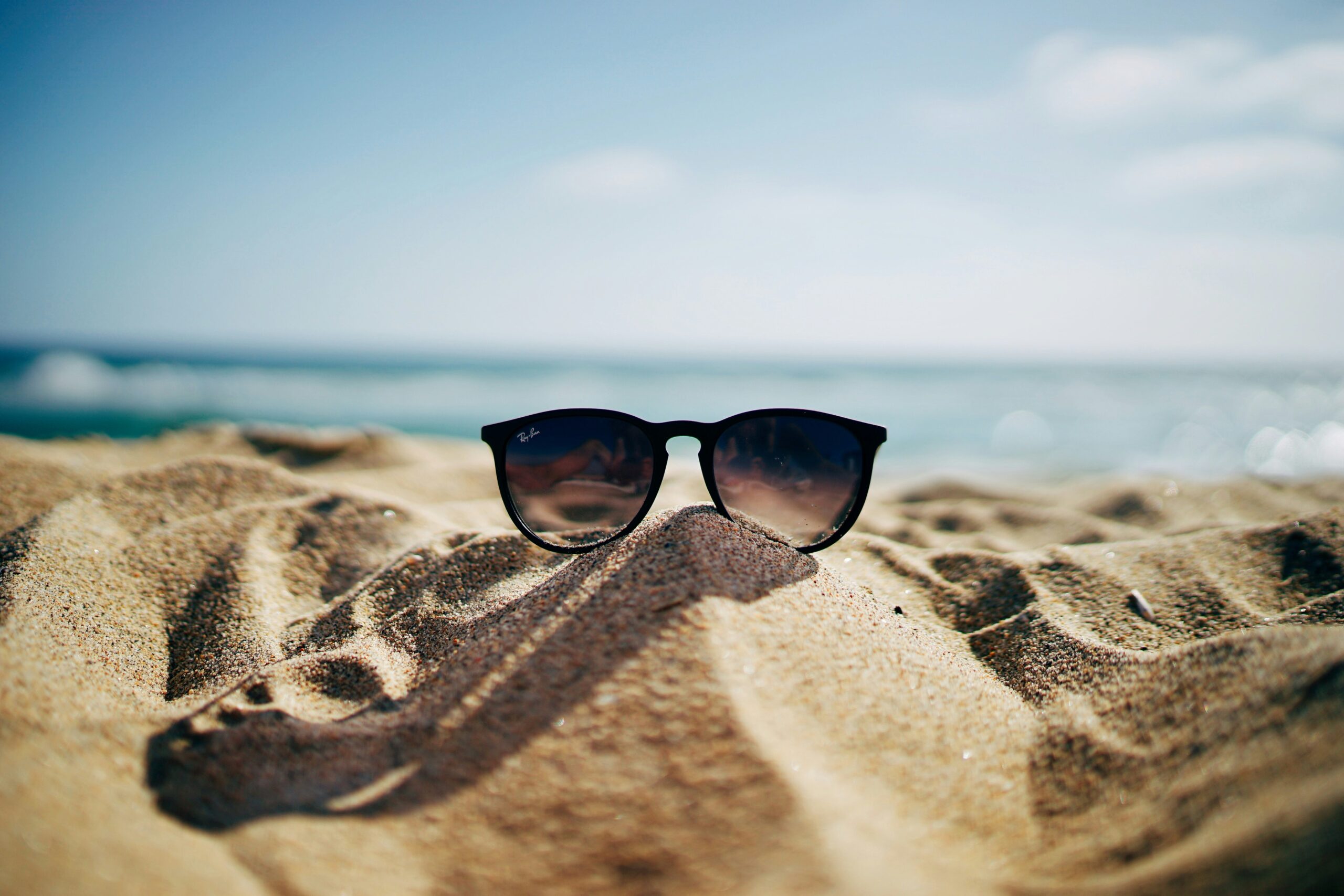 A pair of black sunglasses resting on a sand dune with ocean waves in the background.
