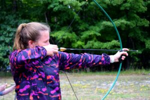 A focused player draws an arrow in an outdoor setting, demonstrating skill and concentration during an archery or arrow tag match. - Brainy Actz Escape Rooms A focused player draws an arrow in an outdoor setting, demonstrating skill and concentration during an archery or arrow tag match.