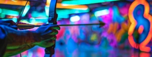 34a140ef 48af 4b68 9067 b3d0e6fe80d8webp - Brainy Actz Escape Rooms Realistic close-up of a player's hand holding a recurve bow with a foam-tipped arrow, ready to shoot in an indoor archery tag arena. The background has colorful graffiti walls and glowing lights. The person's arm is clearly visible. The image is focused on the bow and arrow with a blurry background. The bow can be green or black, and the arrow should have a soft foam tip for safety.