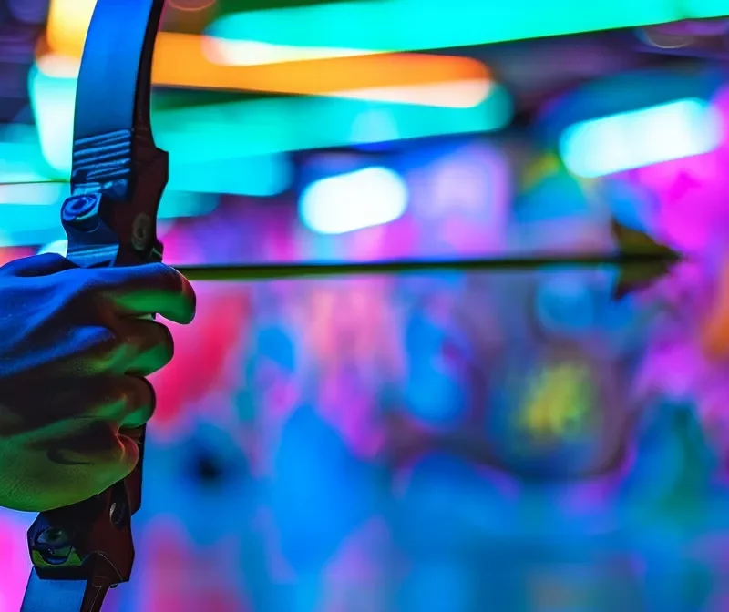 Realistic close-up of a player's hand holding a recurve bow with a foam-tipped arrow, ready to shoot in an indoor archery tag arena. The background has colorful graffiti walls and glowing lights. The person's arm is clearly visible. The image is focused on the bow and arrow with a blurry background. The bow can be green or black, and the arrow should have a soft foam tip for safety.