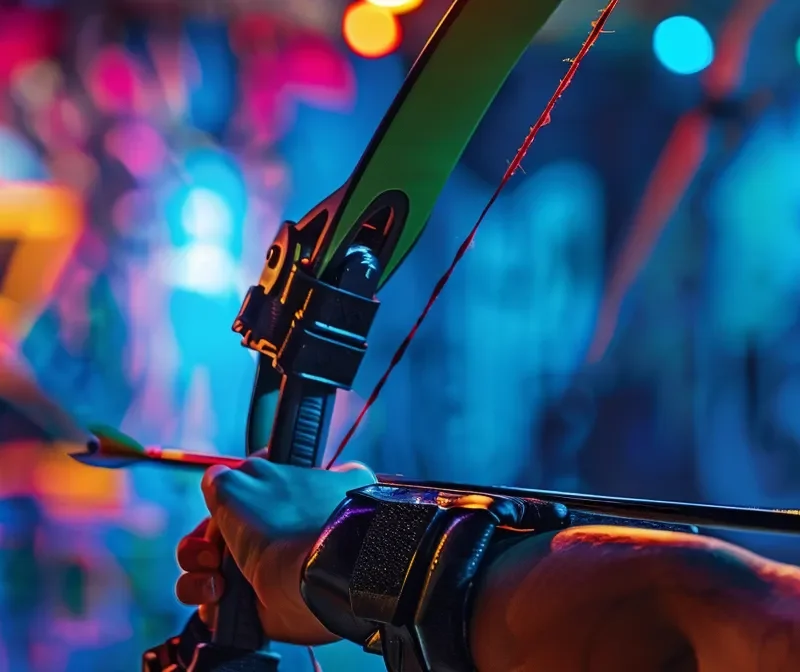 Realistic close-up of a player's hand holding a recurve bow with a foam-tipped arrow, ready to shoot in an indoor archery tag arena. The background has colorful graffiti walls and glowing lights. The person's arm is clearly visible. The image is focused on the bow and arrow with a blurry background. The bow can be green or black, and the arrow should have a soft foam tip for safety.