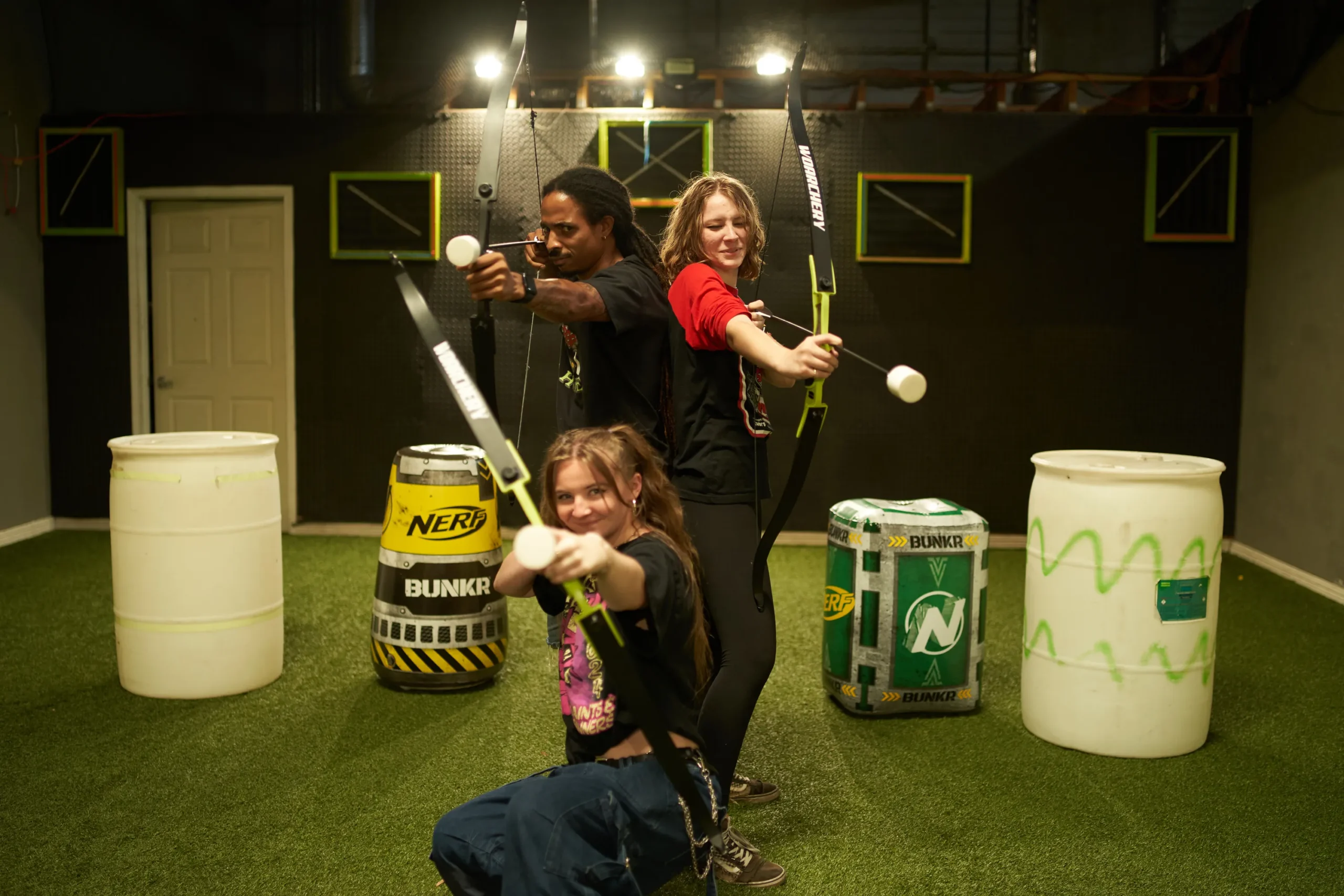 Three players pose with foam-tipped arrows in a well-padded indoor arrow tag arena, highlighting safe venue design and setup. - Brainy Actz Escape Rooms Three players pose with foam-tipped arrows in a well-padded indoor arrow tag arena, highlighting safe venue design and setup.