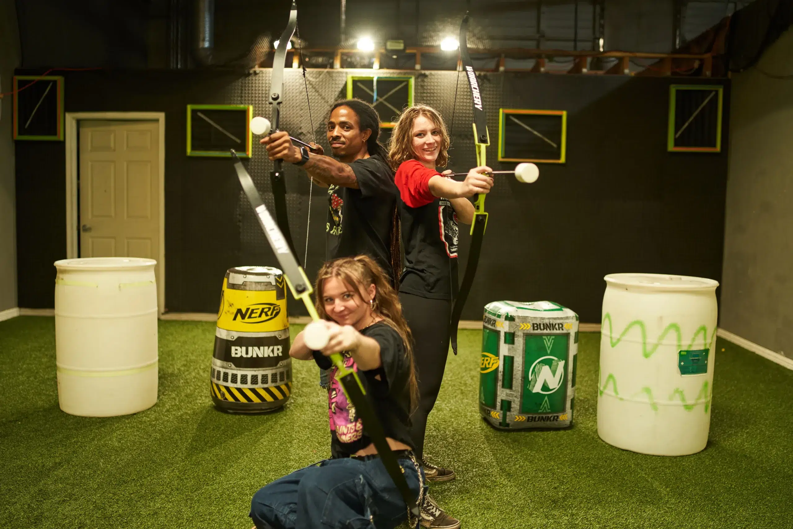 Three players pose confidently with foam-tipped arrows and bows in an indoor arena, illustrating a fun and safe archery arrow tag experience commonly asked about in rental FAQs. - Brainy Actz Escape Rooms Three players pose confidently with foam-tipped arrows and bows in an indoor arena, illustrating a fun and safe archery arrow tag experience commonly asked about in rental FAQs.