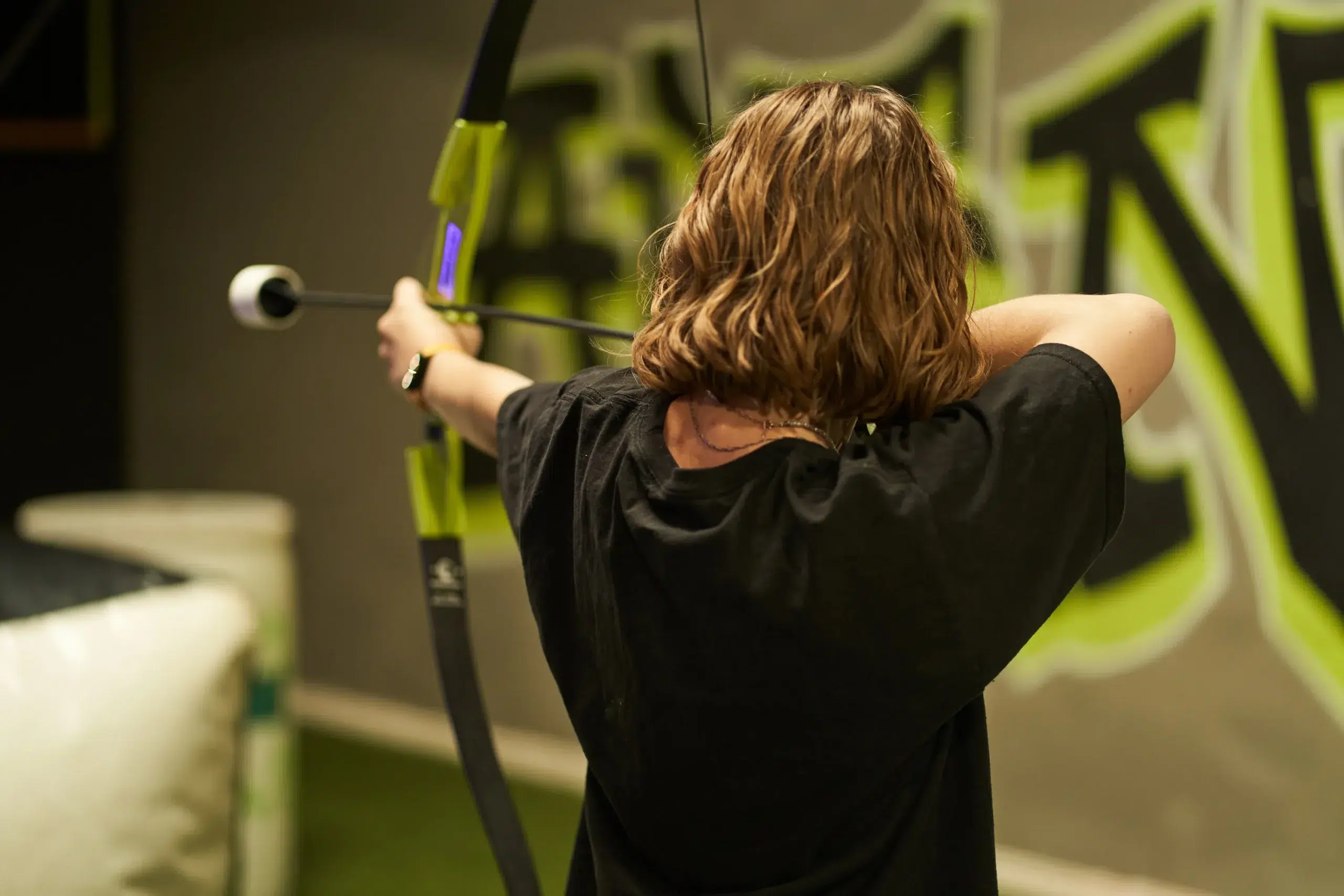 A player aims a foam-tipped arrow during an indoor archery tag match, illustrating gameplay without full-face protection but highlighting optional safety gear. - Brainy Actz Escape Rooms A player aims a foam-tipped arrow during an indoor archery tag match, illustrating gameplay without full-face protection but highlighting optional safety gear.