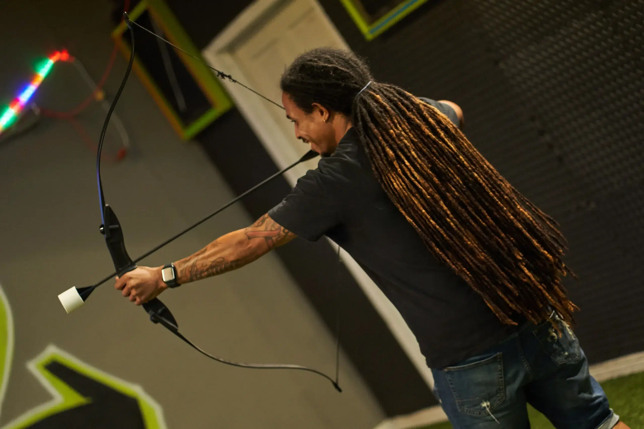 A player draws a bow with a foam-tipped arrow in an indoor arena, emphasizing the importance of following safety protocols in archery arrow tag equipment rentals. - Brainy Actz Escape Rooms A player draws a bow with a foam-tipped arrow in an indoor arena, emphasizing the importance of following safety protocols in archery arrow tag equipment rentals.