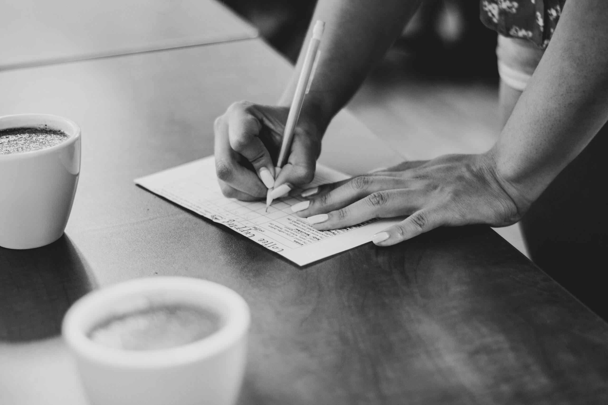 A person writes on a score sheet at a table with coffee cups nearby, representing the process of tracking and recording scores. - Brainy Actz Escape Rooms A person writes on a score sheet at a table with coffee cups nearby, representing the process of tracking and recording scores.