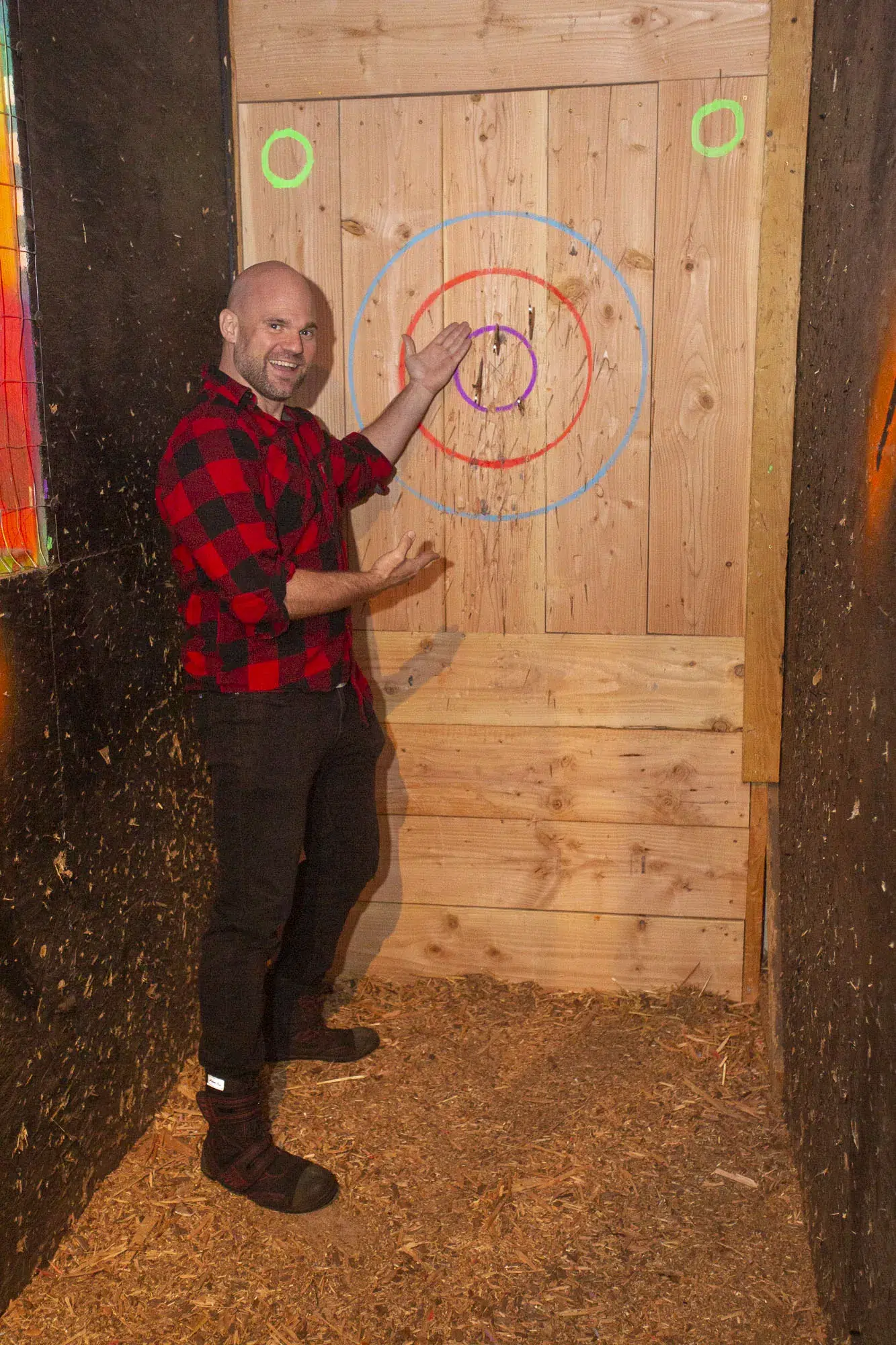A smiling participant points at a bullseye on a wooden target board, highlighting the fun and camaraderie that group engagement brings to archery tag activities. - Brainy Actz Escape Rooms A smiling participant points at a bullseye on a wooden target board, highlighting the fun and camaraderie that group engagement brings to archery tag activities.