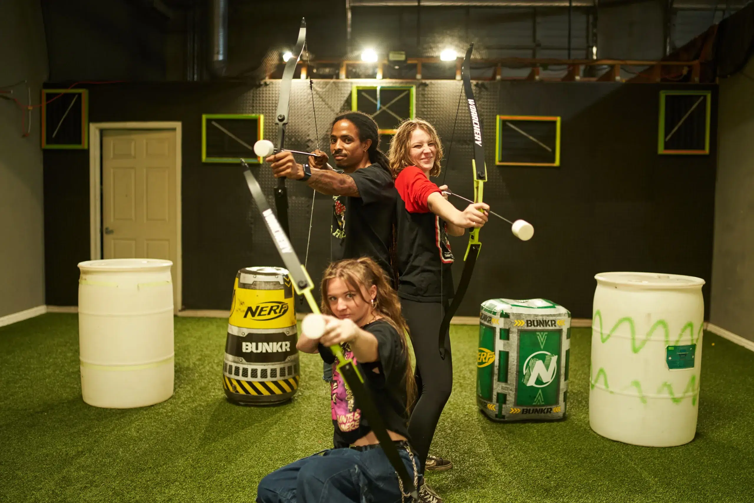 Three players pose with bows drawn in a clearly marked arrow tag arena, highlighting the importance of designated game zones for organized, chaos-free play. - Brainy Actz Escape Rooms Three players pose with bows drawn in a clearly marked arrow tag arena, highlighting the importance of designated game zones for organized, chaos-free play.