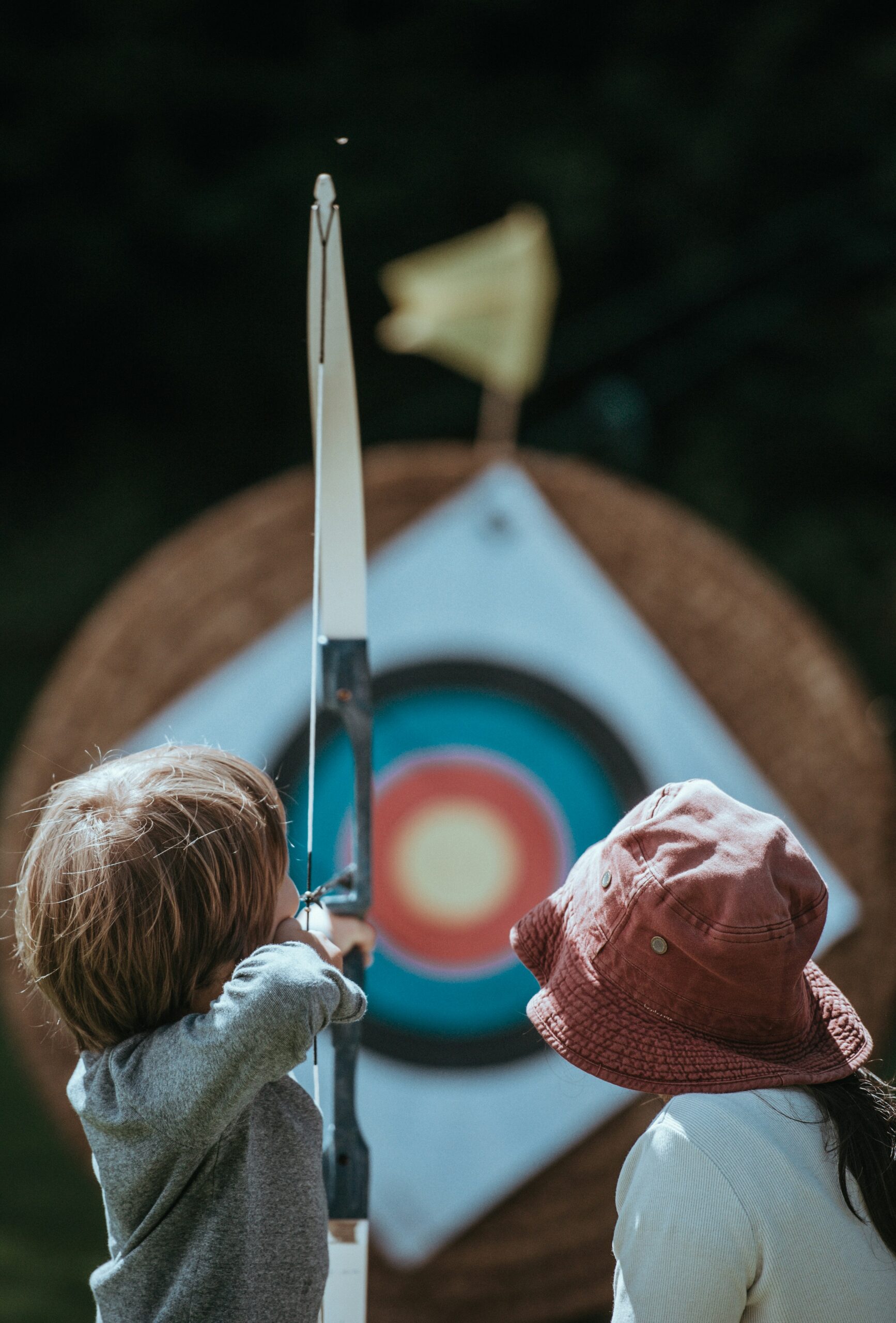 A young child practices archery with adult supervision near a target, reflecting the importance of adapting rules to ensure safety for younger players in arrow tag. - Brainy Actz Escape Rooms A young child practices archery with adult supervision near a target, reflecting the importance of adapting rules to ensure safety for younger players in arrow tag.