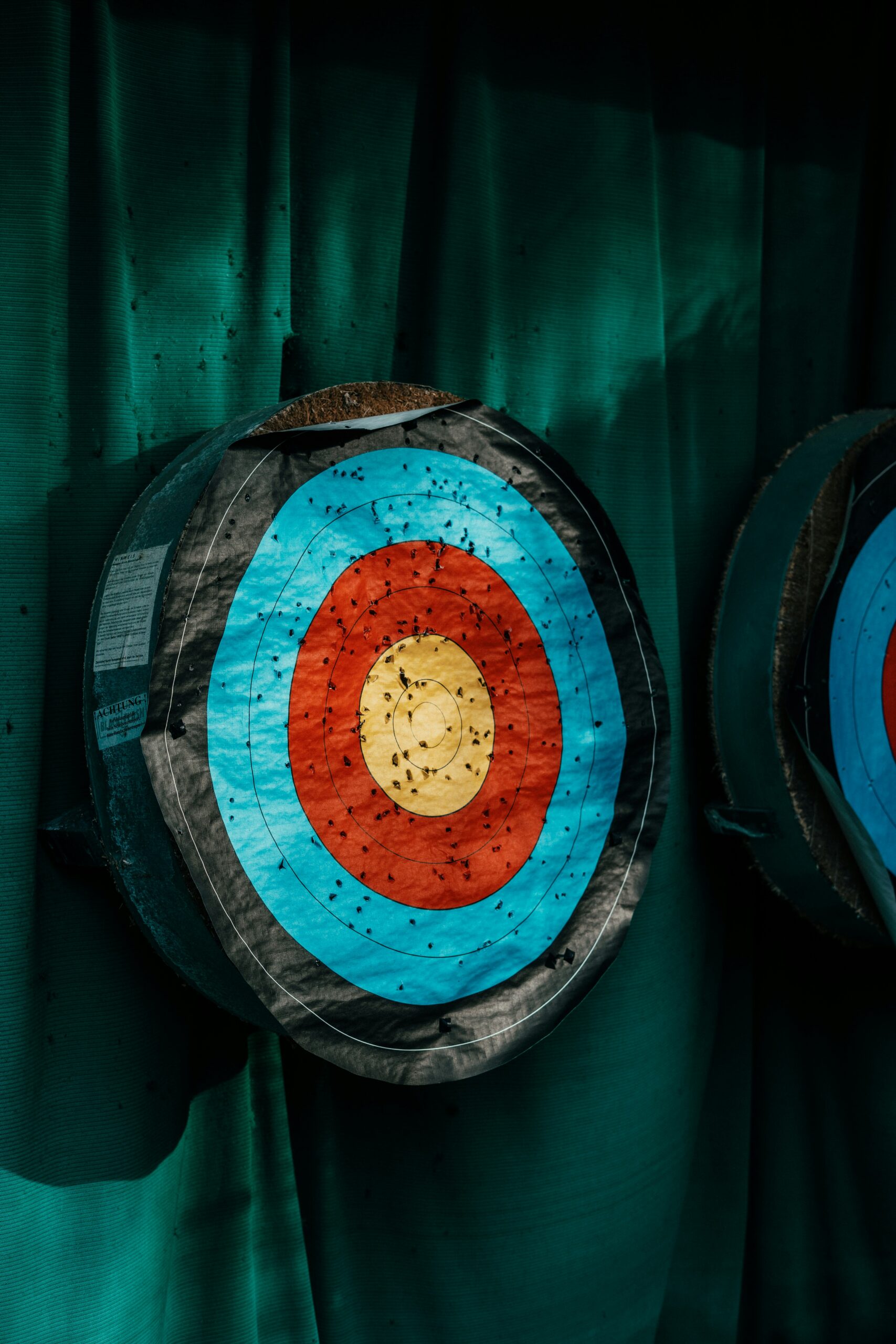 A well-worn bullseye target with layered foam backing and colorful scoring rings demonstrates a practical and effective approach to building custom or DIY Archery Tag targets. - Brainy Actz Escape Rooms A close-up of a layered bullseye-style target riddled with impact marks, illustrating the durability and design considerations for building effective custom Archery Tag targets.