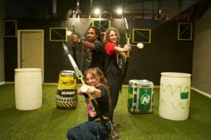 Three archery tag players pose with drawn bows in an indoor arena, showcasing how regular participation in archery activities boosts stamina and physical endurance. - Brainy Actz Escape Rooms Three archery tag players pose with drawn bows in an indoor arena, showcasing how regular participation in archery activities boosts stamina and physical endurance.
