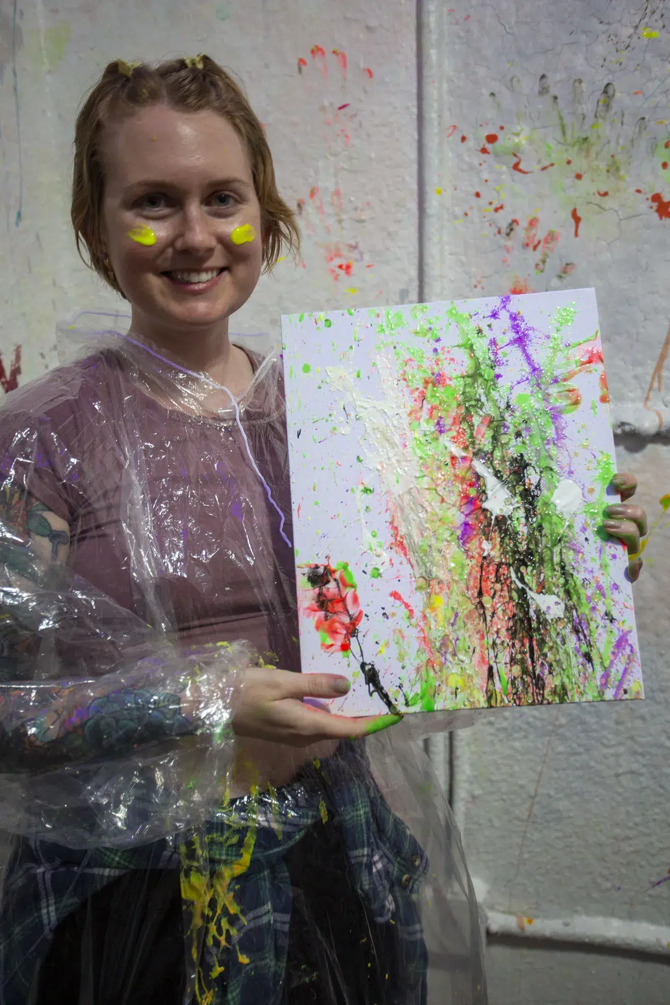 A person wearing a plastic poncho smiles while holding a splatter-painted canvas created during a fun beginner-friendly painting session.