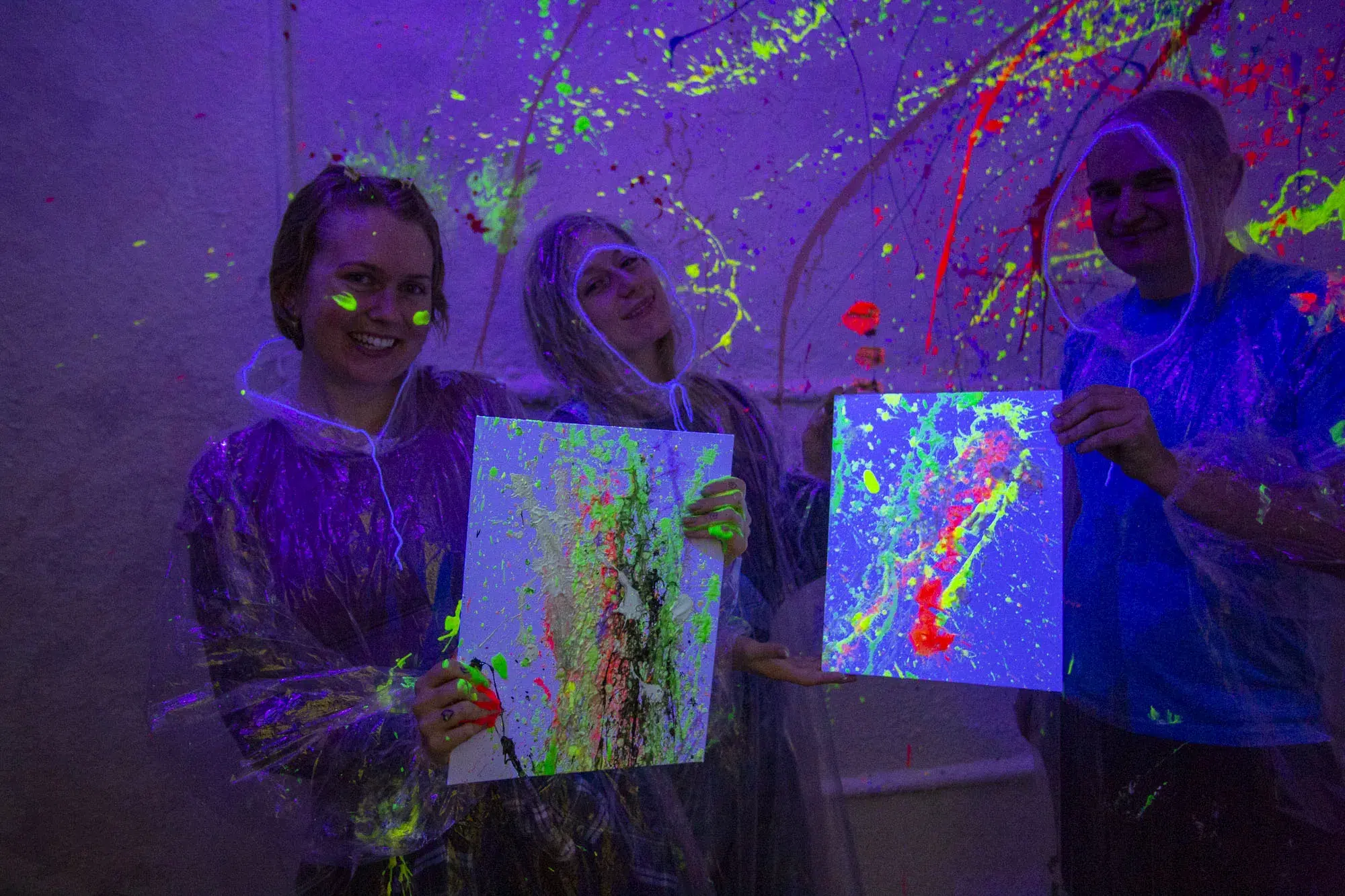 Group of friends in plastic ponchos proudly display their neon splatter art in a glow-in-the-dark room, perfect for lively group celebrations or team events.