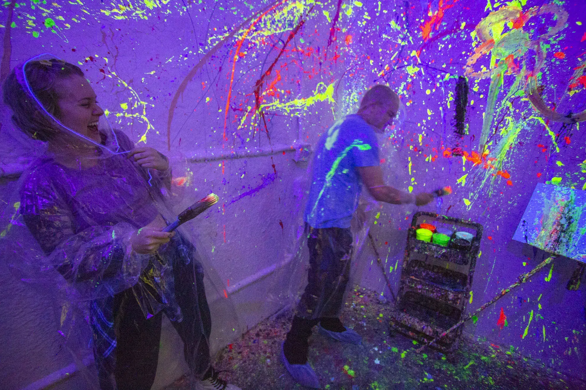 Two people laugh and paint together in a neon-lit splatter room, showing how this vibrant, creative activity brings groups closer through shared fun.