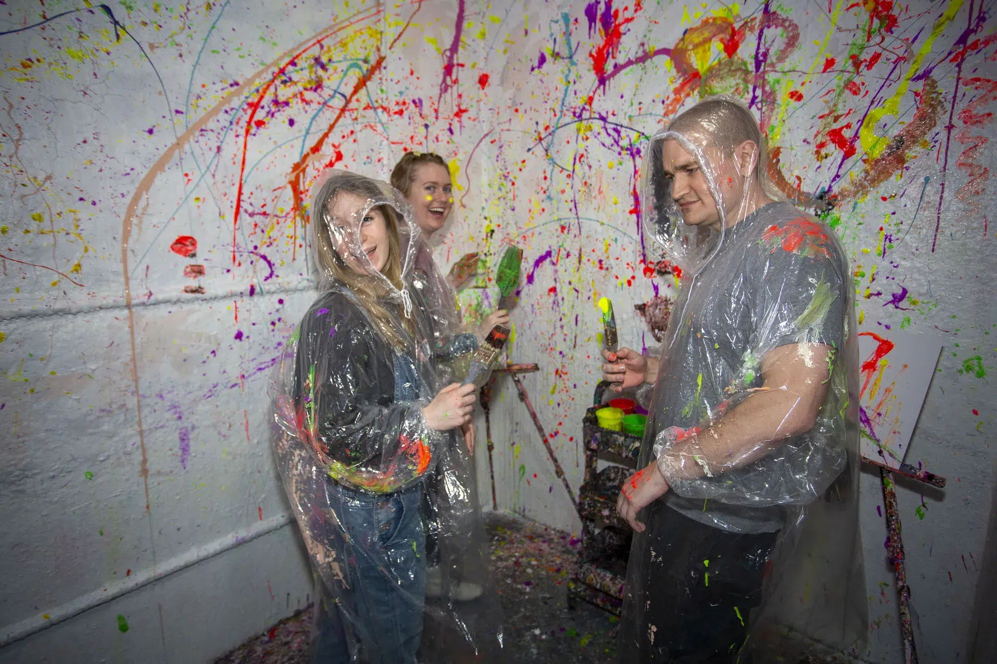 Group of people in clear ponchos holding paintbrushes in a splatter paint room, ready for a fun and safe Brainy Actz session.