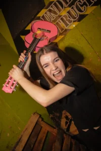 A woman in a wreck room smashing a pink guitar with a wild smile, capturing the freedom of a rage session for any mood. - Brainy Actz Escape Rooms A woman in a wreck room smashing a pink guitar with a wild smile, capturing the freedom of a rage session for any mood.