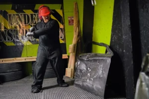 A person in full protective gear swings a bat at metal debris in a rage room, demonstrating how to smash objects safely and responsibly. - Brainy Actz Escape Rooms A person in full protective gear swings a bat at metal debris in a rage room, demonstrating how to smash objects safely and responsibly.