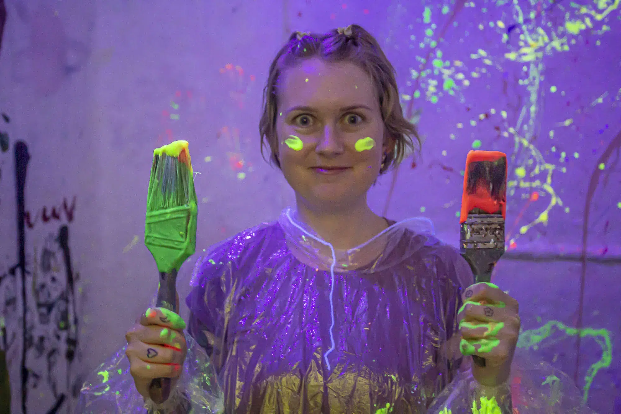 Guest in a paint-covered poncho grins while holding neon-glowing paintbrushes, capturing the playful and hands-on fun of a splatter paint room experience.