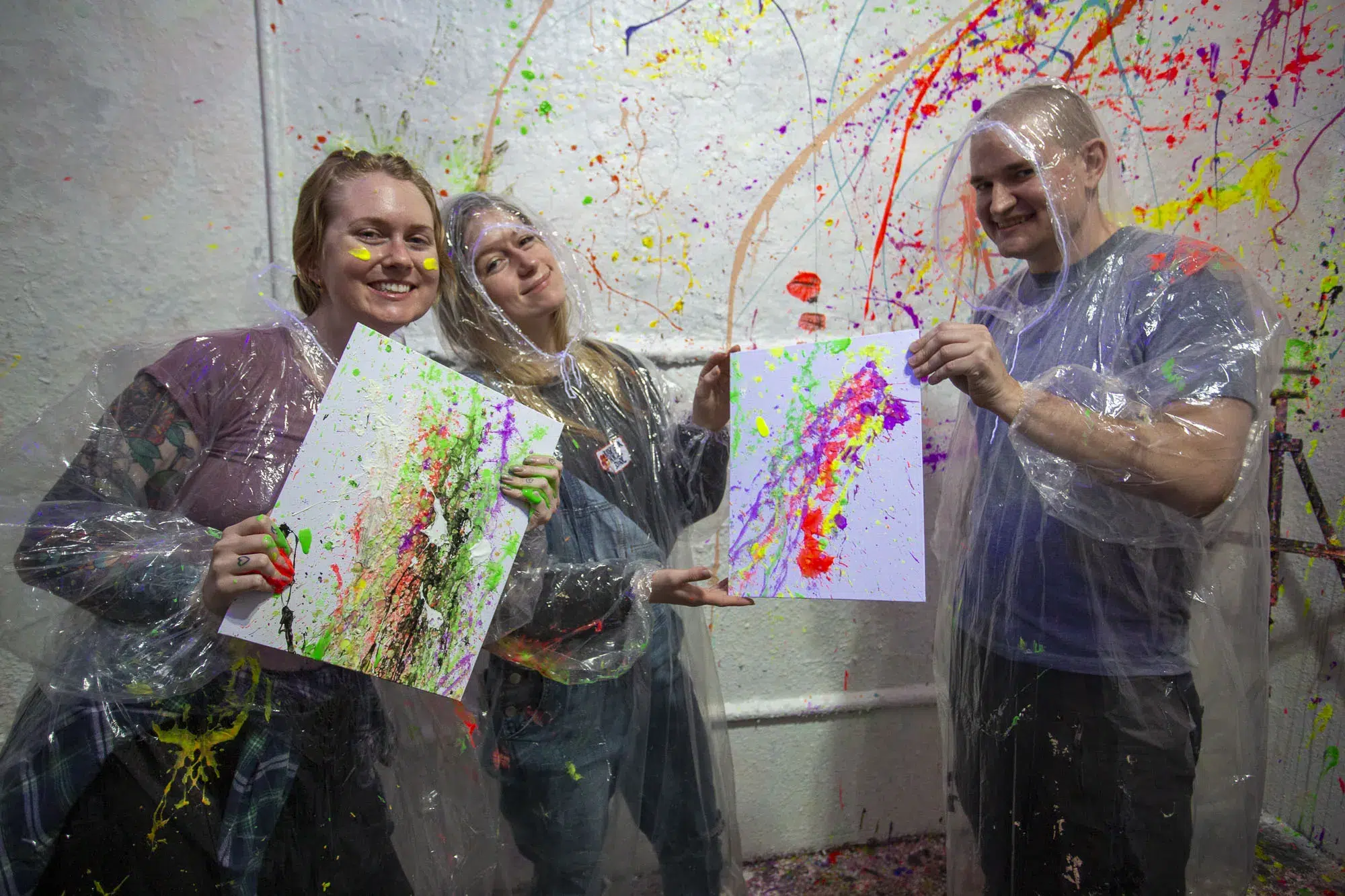 Three people in ponchos proudly display their splatter art on canvas, smiling in a colorful, paint-covered room after a fun team activity.